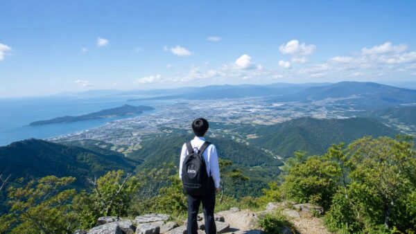 制服姿の男子高校生が、初めて登った低山の頂上で清々しい表情で景色を眺めている