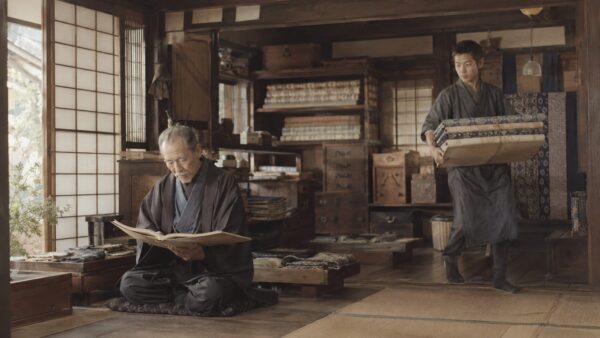 明治〜大正期の商家の店内で、主人が帳簿を見ながら、若い丁稚が荷物を運んでいる